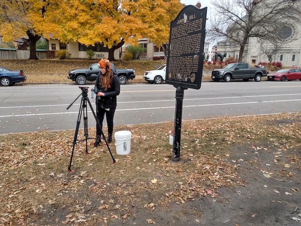 Woman standing on boulevard holding video camera with camera tripod in front of her.  Next to her is black cast aluminum historical marker.