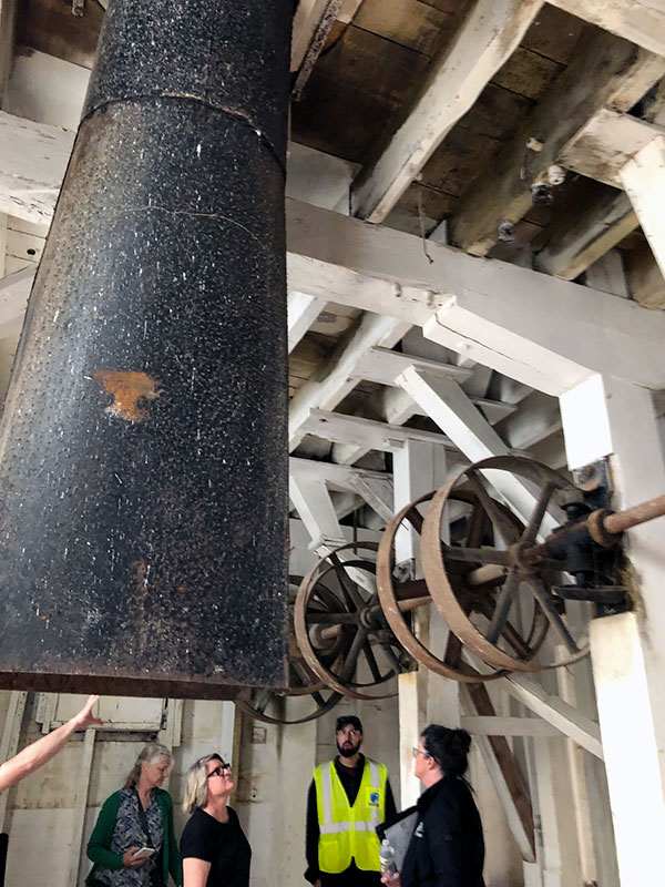 Four people looking at milling equipment above them. Long dark shaft in above foreground along with large wheel gears.