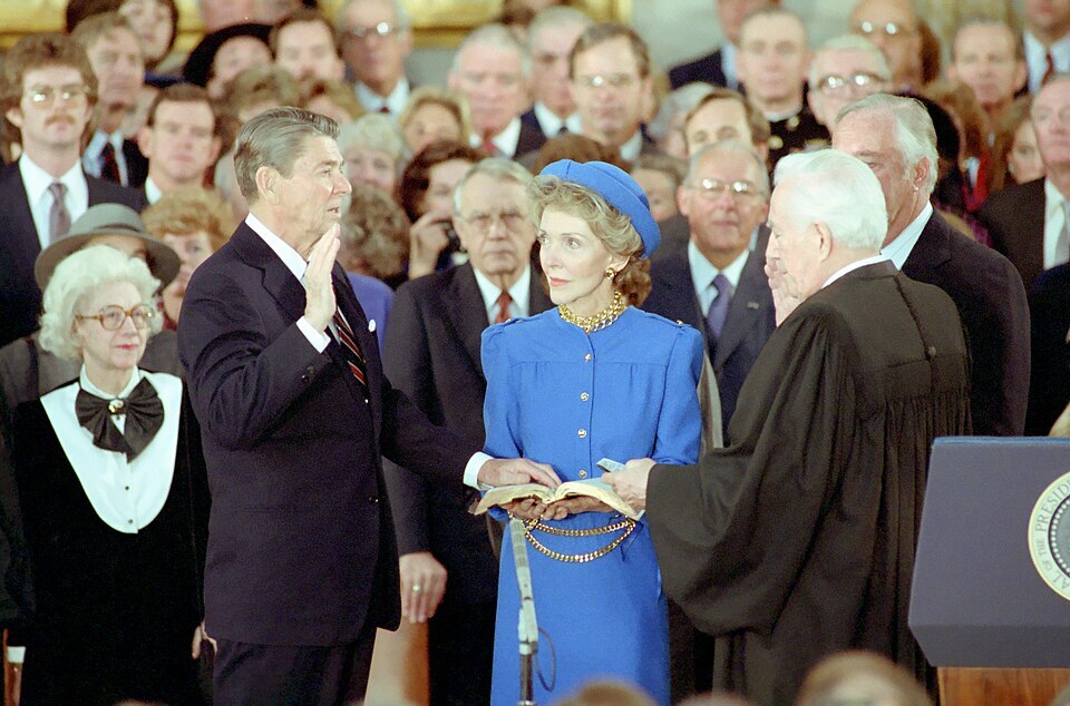 Three people stand in a flattened triangular formation in front of a crowd of spectators. At left, a man raises his right hand to mirror the man holding up his own right hand across from him. A woman stands in the middle, wearing a blue dress and looking on.