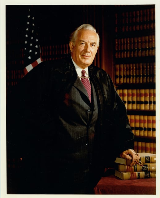 A white-haired man stands next to a desk with three books on it. He wears the robe of a Supreme Court justice. Behind him area an American flag and a bookshelf.