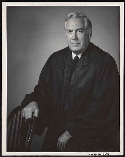 A man in a black robe sits next to a chair, with his right hand resting on it.