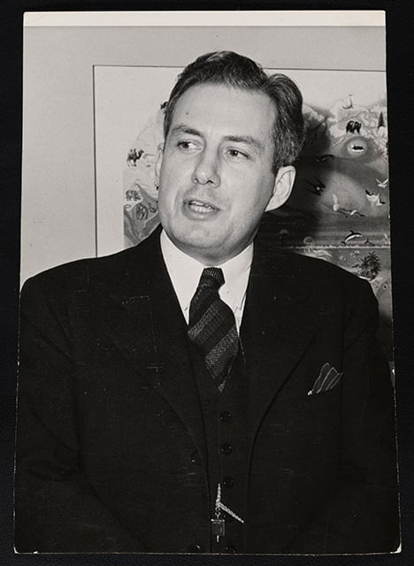 A standing man wearing a suit and tie, shown from the waist up to his head. Behind him is a piece of art hanging on a wall. 