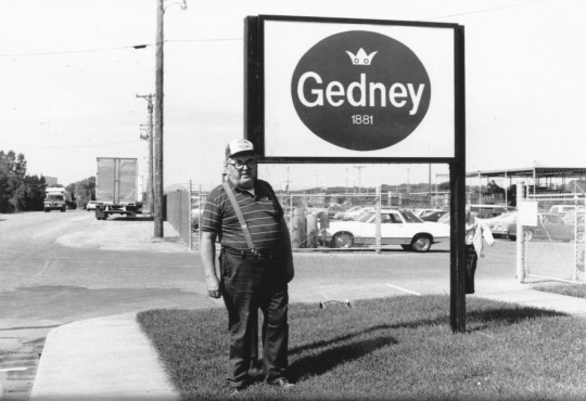 Black and white photograph of a man standing to the left of the Gedney sign, outside the Chaska factory, c.1980s.